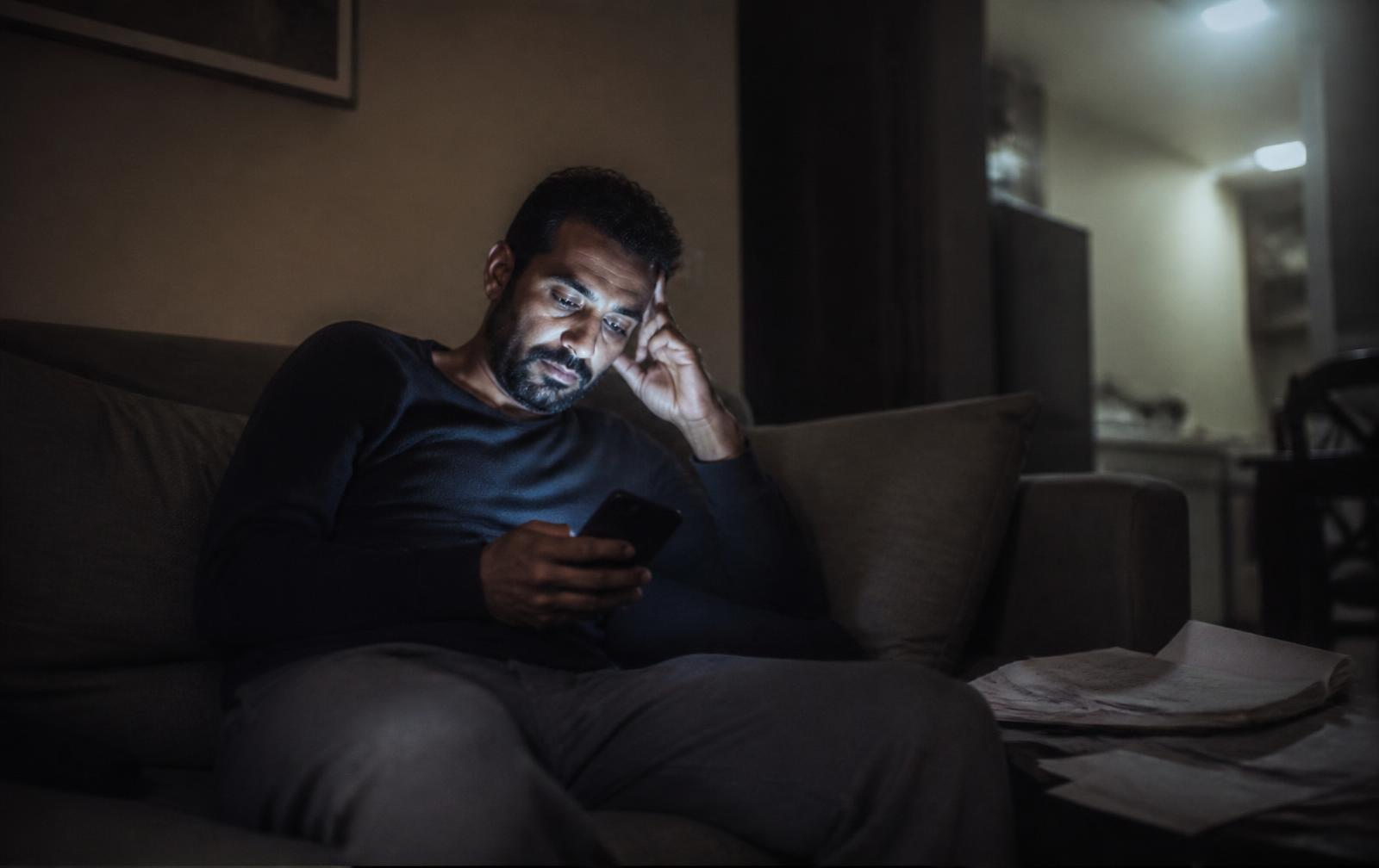 A father alone on a sofa late at night, lit only by his phone, papers beside him