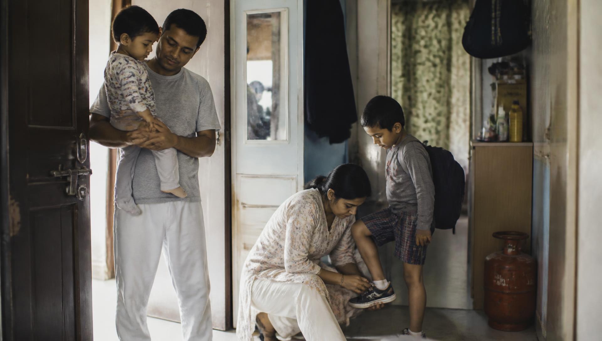 A family in a doorway on a weekday morning, one parent holding a toddler while the other laces a child's shoes