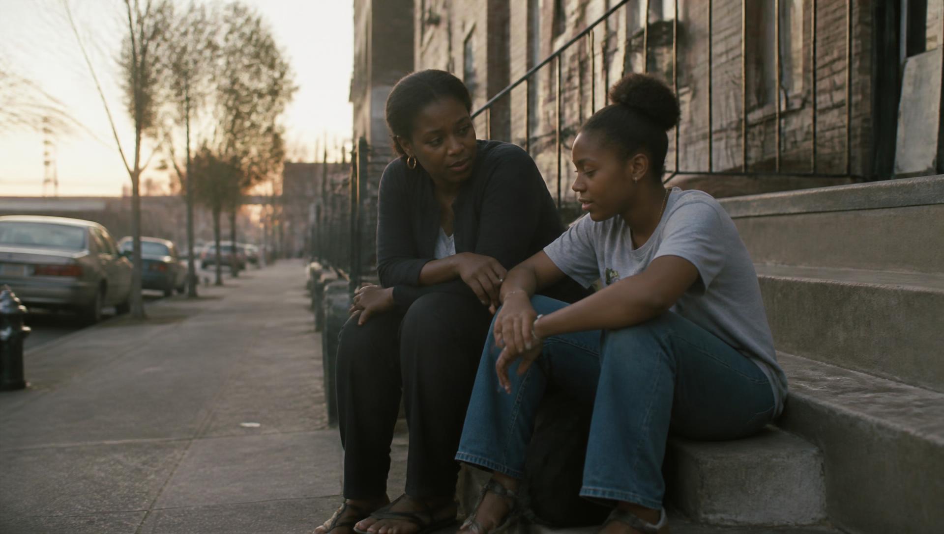 A mother and her teenage daughter sitting on the steps outside their apartment building at dusk, talking