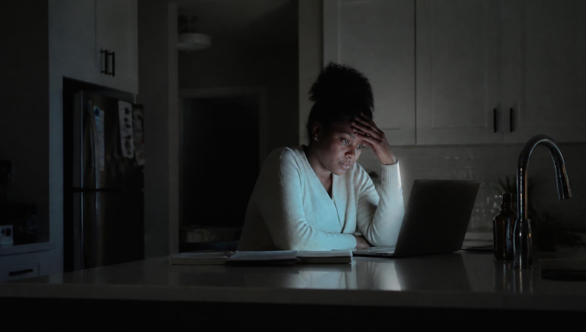 A parent alone at a kitchen counter late at night, lit only by a laptop